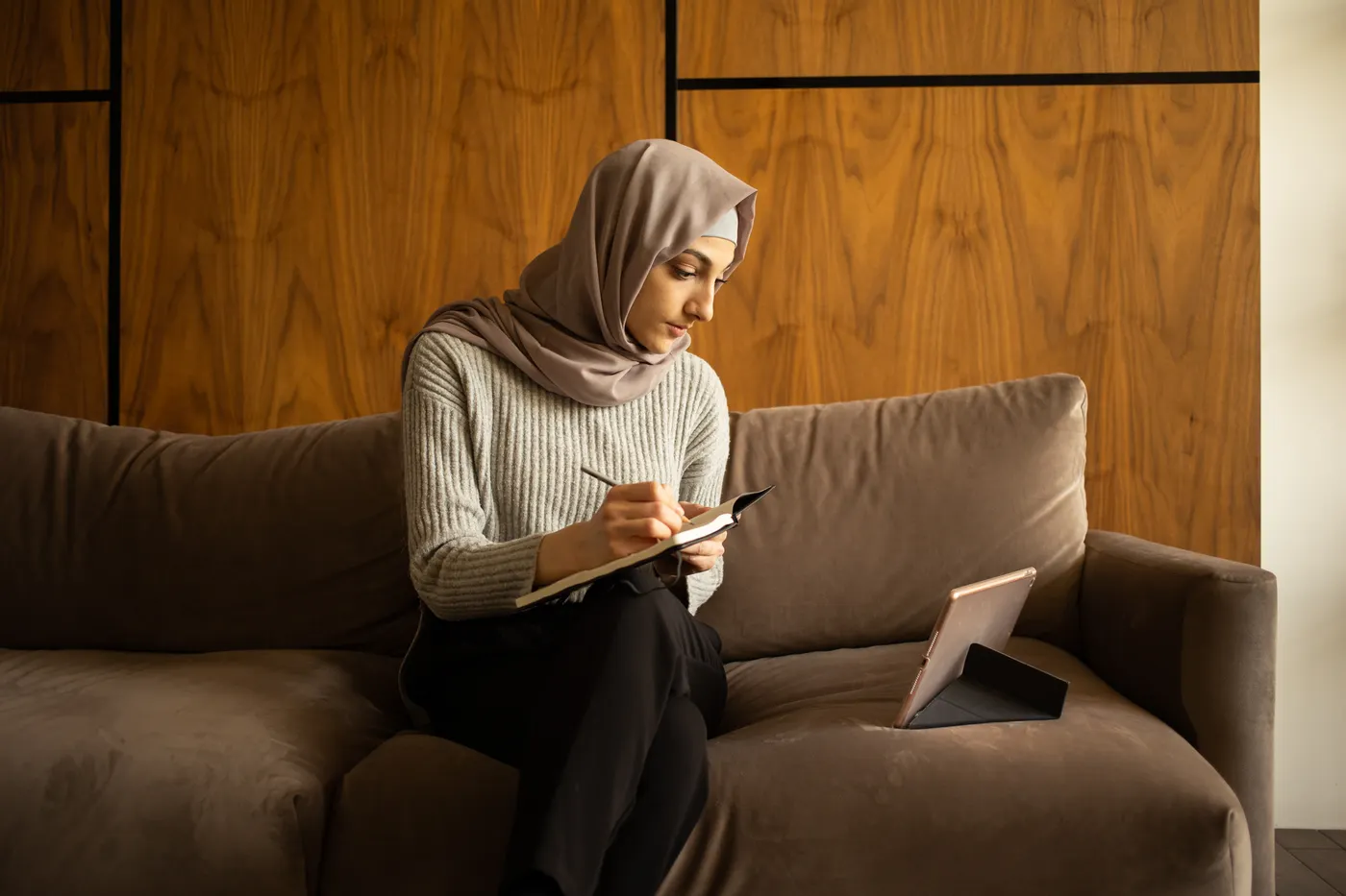 A student takes notes on a sofa with a tablet open beside her